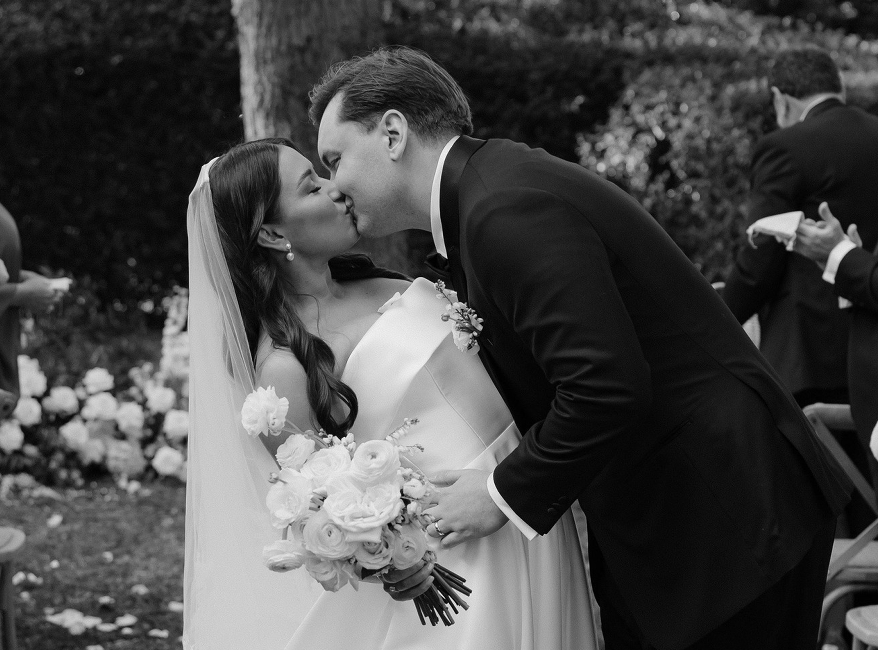 A bride and groom share a kiss outdoors during their wedding, surrounded by flowers and greenery. The bride holds a bouquet of white roses and wears a veil, while the groom is in a dark suit. The scene is intimate and romantic.