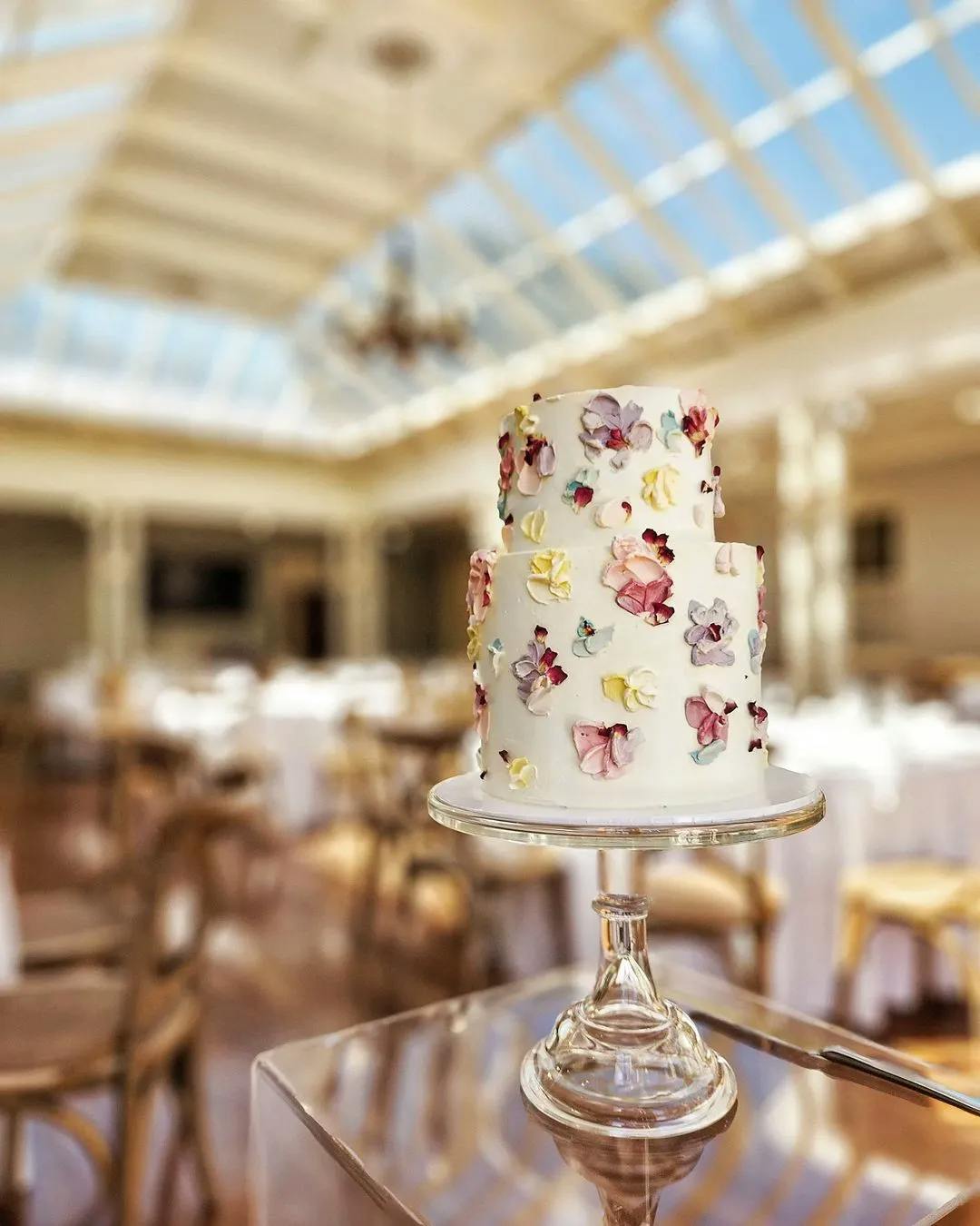 A two-tiered white wedding cake decorated with colorful flower petals is displayed on a glass stand. The background features a bright, elegant venue with wooden chairs and round tables covered in white tablecloths under a skylight ceiling.