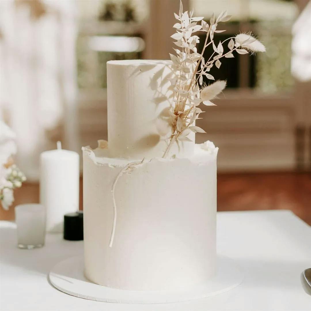 A two-tier white wedding cake decorated with minimal white floral elements and delicate, dried grass. The cake is placed on a white tablecloth with blurred background elements, enhancing its elegant simplicity.