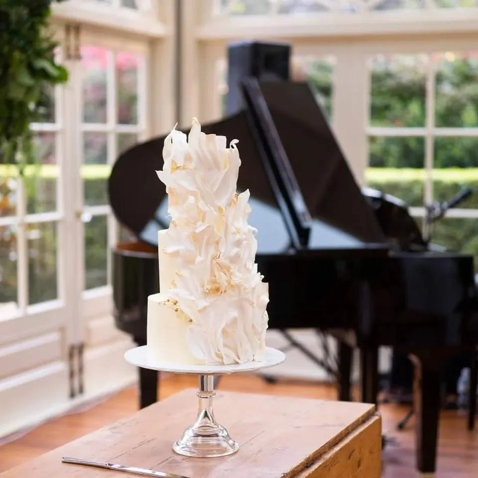 A two-tiered, white wedding cake adorned with delicate, vertical floral decorations, displayed on a clear cake stand. The cake is set in an elegant room with large windows, natural light, a grand piano, and lush greenery in the background.