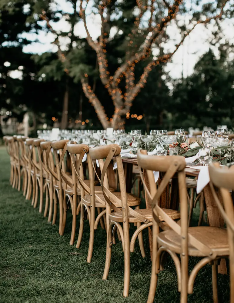 A long outdoor dining table is set with elegant place settings, including wine glasses and plates, surrounded by wooden chairs. The background features a tree wrapped in twinkling lights, creating a warm and inviting atmosphere amidst a grassy area.