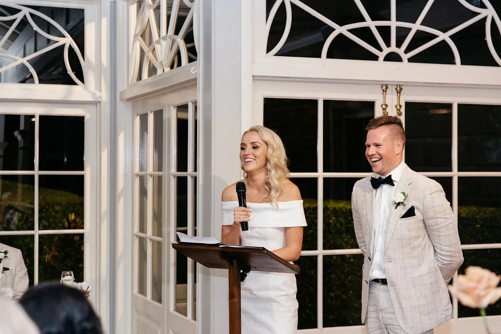 A woman in a white off-shoulder dress is giving a speech at a wooden podium, holding a microphone, and smiling. A man in a light-colored suit with a black bow tie stands next to her, also smiling. They are indoors near large windows with decorative panes.