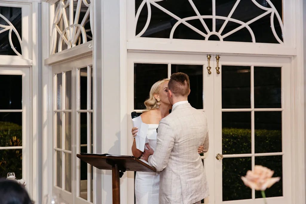 A couple stands in front of large glass doors with intricate window designs, sharing a kiss. The woman, holding a microphone, wears a white dress, while the man in a light-colored suit embraces her. The scene is warmly lit, suggesting an intimate moment or celebration.