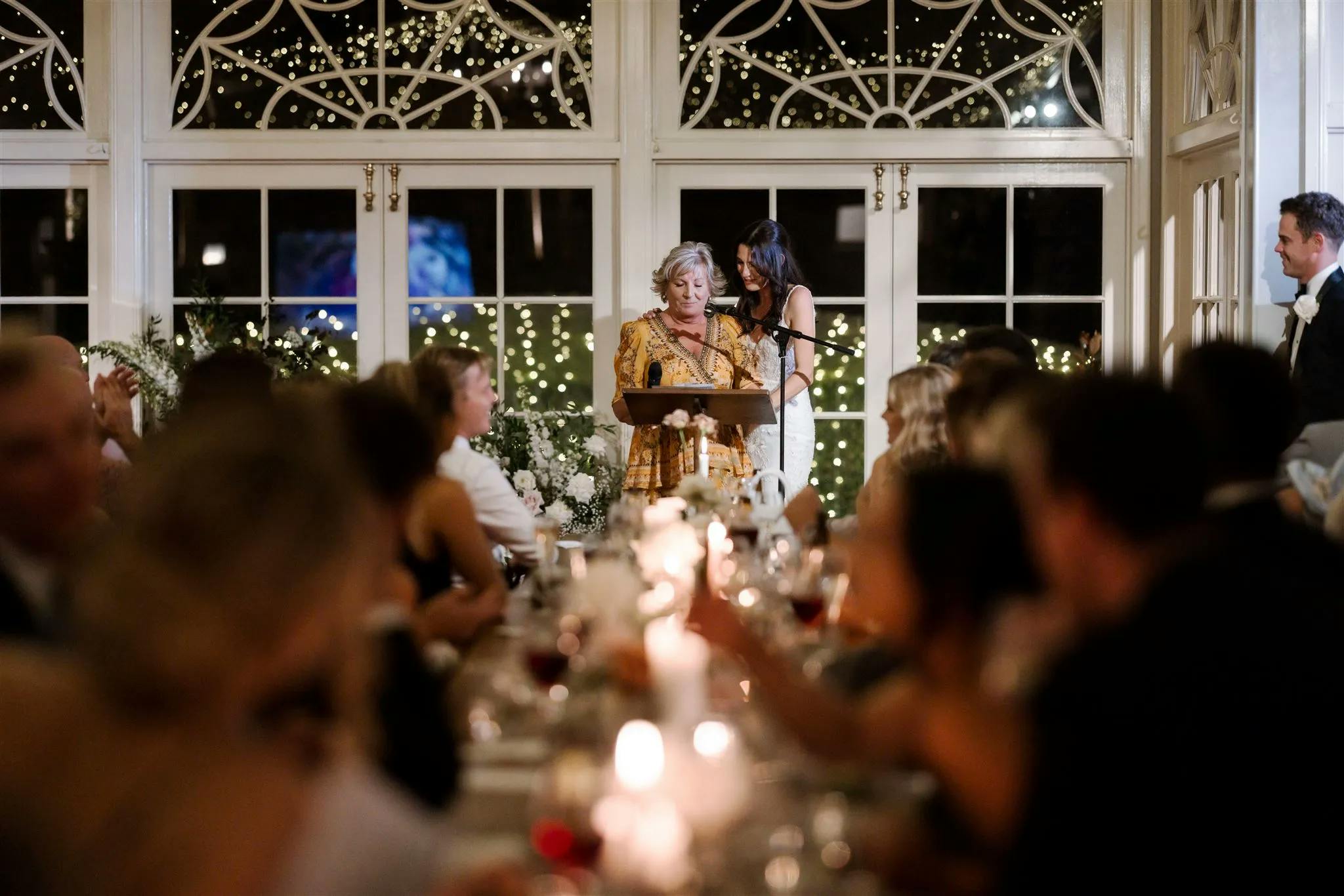 A woman in a gold and black dress stands at a podium delivering a speech, while a second woman in a white dress stands beside her. Guests are seated at dimly lit tables in a decorated venue with fairy lights and arched windows in the background.