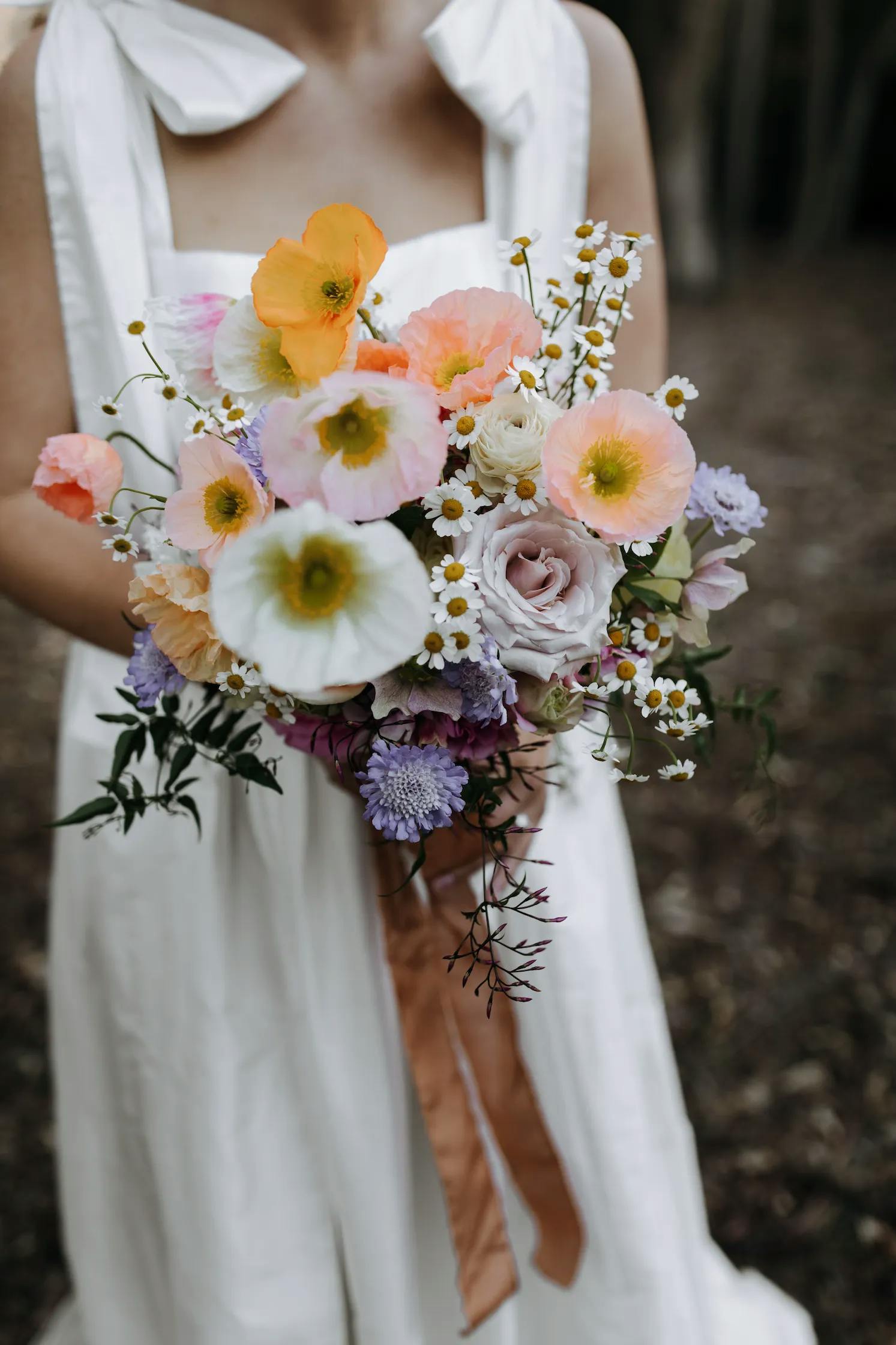 A person wearing a sleeveless white dress holds a vibrant bouquet of flowers, including orange poppies, white daisies, and light purple blooms, with a brown ribbon tied around the stems. The background is outdoors with a blurred, earthy ground.