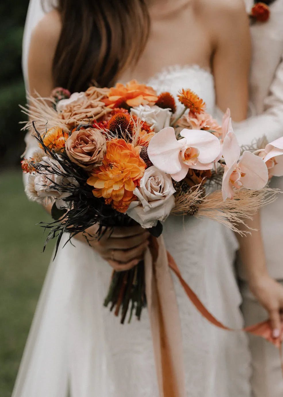 A bride in a strapless white wedding dress holding a vibrant bouquet featuring orange, peach, and white flowers with various greenery. Her partner stands behind her, partially visible, in a light-colored suit. The background is a lush, green outdoor setting.