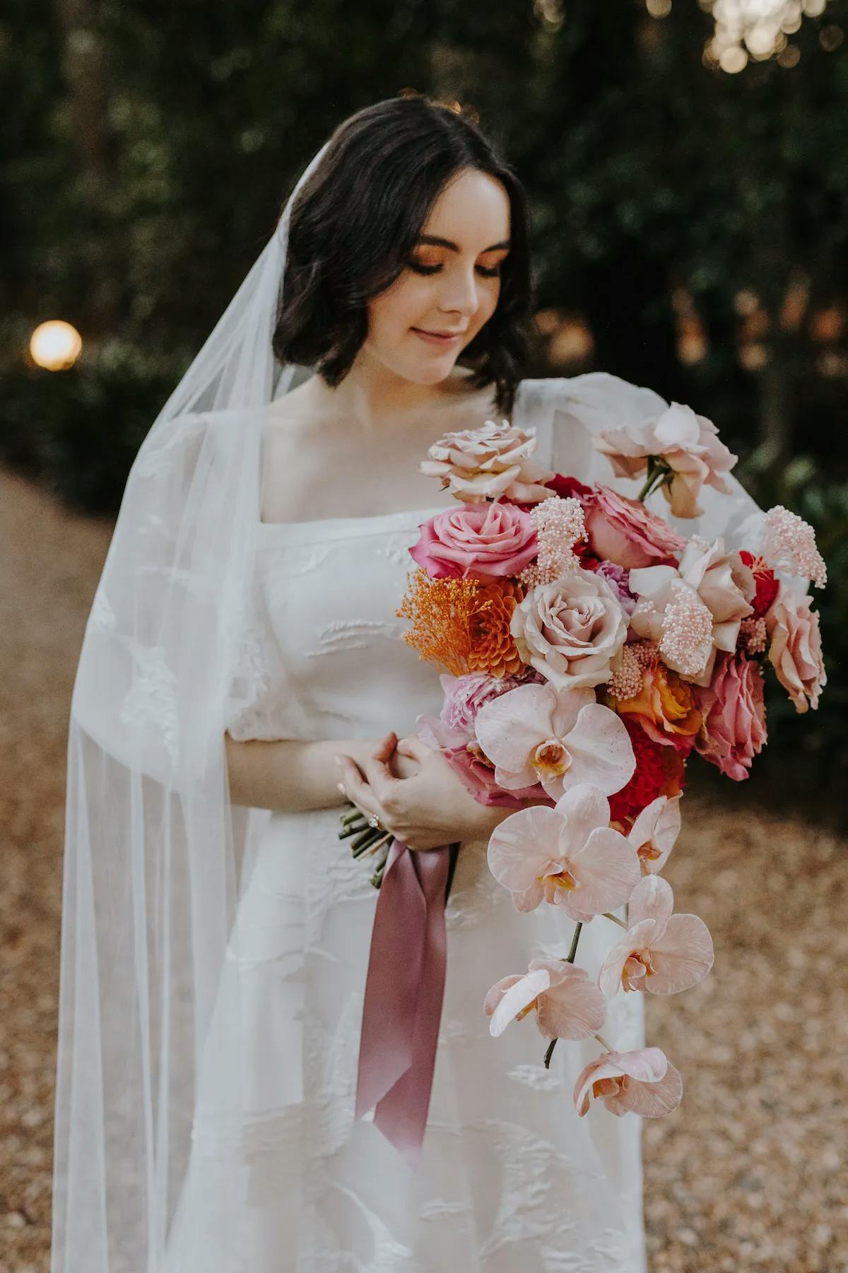 A bride with dark hair looks down while holding a cascading bouquet of pink, peach, and orange flowers. She is wearing a white wedding dress with a sheer veil and stands on a pebble path with greenery in the background, illuminated by soft lighting.