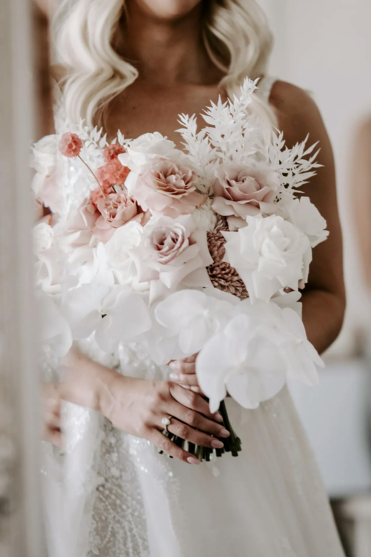 A bride in a wedding dress holds a floral bouquet featuring white orchids, pink roses, and other assorted flowers and greenery. The focus is on the flowers, with the bride's face partially out of frame. The image has a soft, romantic feel.