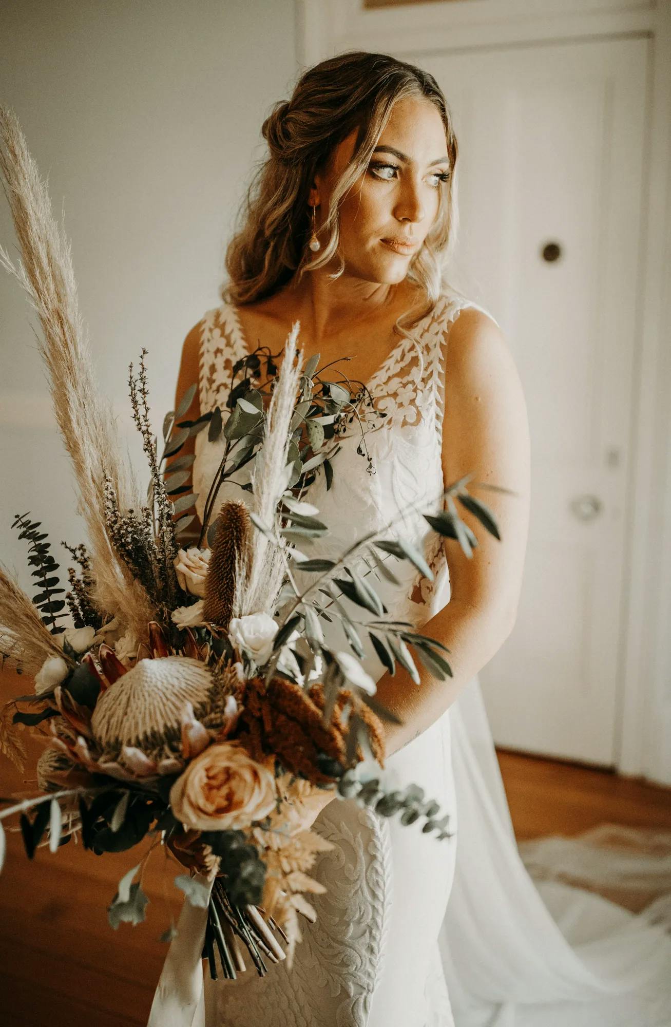 A bride in a lace wedding dress holds a bouquet made of neutral-toned flowers, greenery, and dried elements. She gazes thoughtfully to the side, with soft lighting highlighting her serene expression and the intricate details of her gown and bouquet.