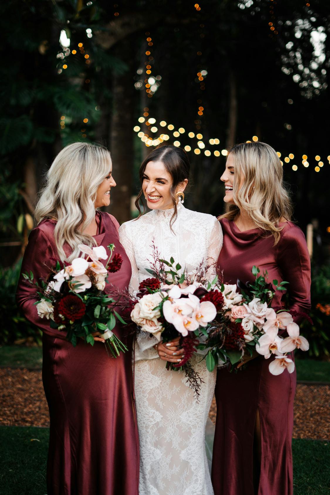 A bride in an elegant white lace dress stands between two bridesmaids in matching burgundy dresses. All three women are smiling joyfully and holding bouquets of flowers, with twinkling string lights and lush greenery in the background.