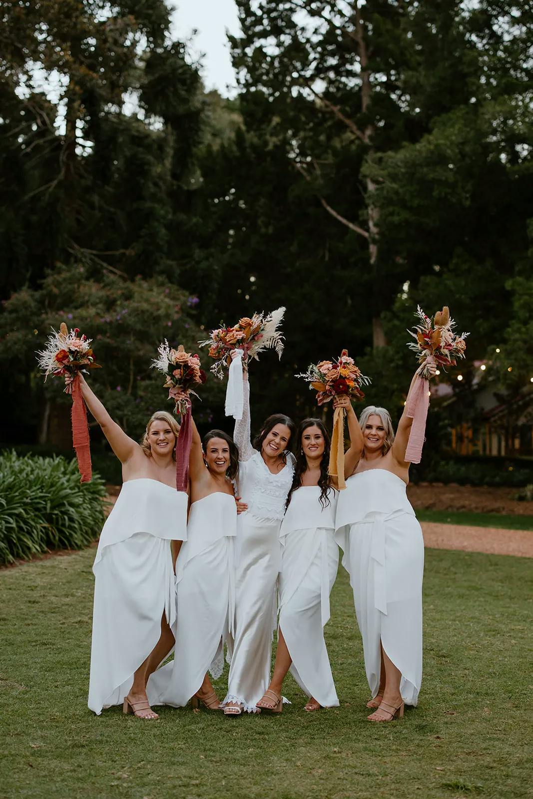 A bride and her four bridesmaids stand on grass outdoors, smiling and celebrating. The bride wears a long white dress, and the bridesmaids wear matching off-shoulder white dresses. Each woman holds a colorful bouquet of flowers, raising them joyfully in the air.