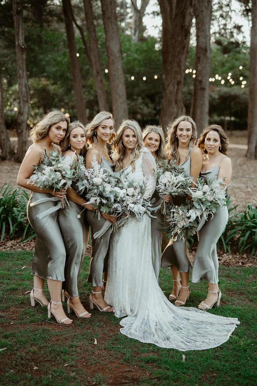A bride in a white lace gown stands with five bridesmaids wearing silver dresses. They are in an outdoor setting with trees and string lights in the background. Each holds a bouquet of flowers, and they are all smiling at the camera.