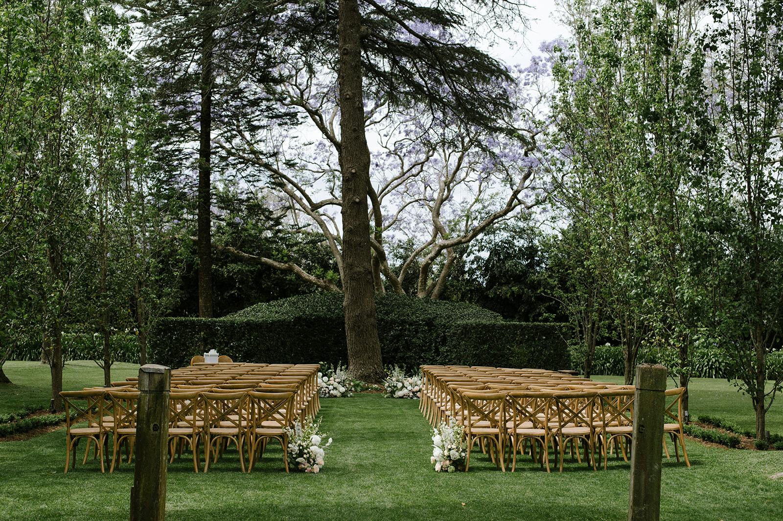 A serene outdoor wedding setup on a lush green lawn. Rows of wooden chairs with floral arrangements on the aisle line a path leading to a large tree at the altar. The area is surrounded by tall trees and greenery, creating an intimate and natural setting.