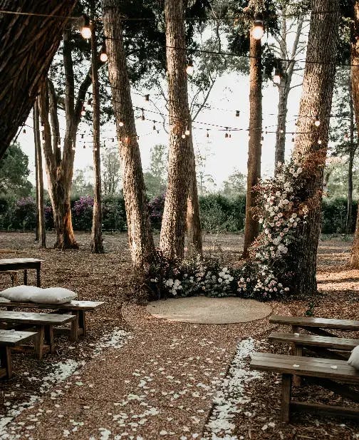 A rustic outdoor wedding setup under tall trees with string lights hanging above. Wooden benches are lined along a petal-strewn aisle leading to an altar area adorned with flowers. The setting conveys a serene, romantic atmosphere in a forest-like environment.
