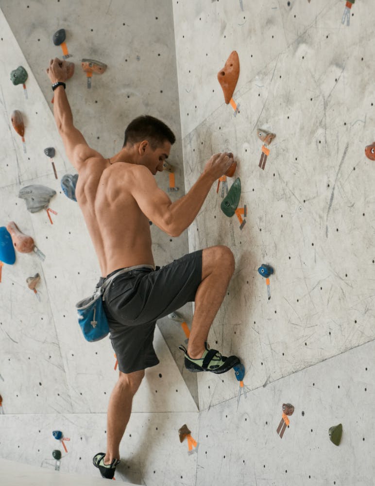 A man is engaged in indoor rock climbing on a wall with variously colored holds. He is shirtless, wearing shorts and climbing shoes with a chalk bag attached to his waist. The wall has an array of handholds and footholds in different shapes and colors.