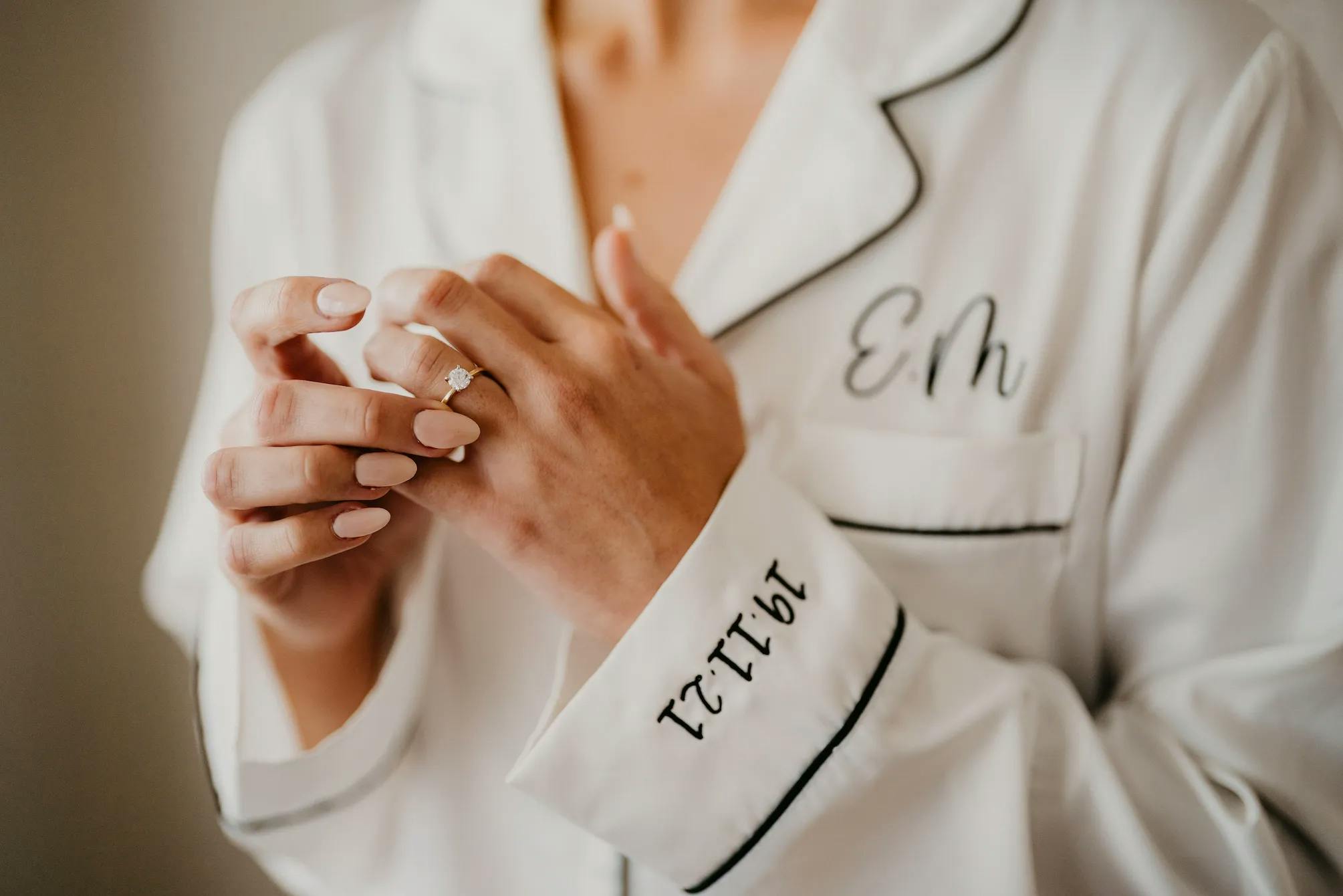 A person wearing a white pajama shirt with black piping, embroidered initials "EM," and the date "12-17-21" on the sleeve. They are displaying a sparkling engagement ring on their left hand and have neatly manicured nails.