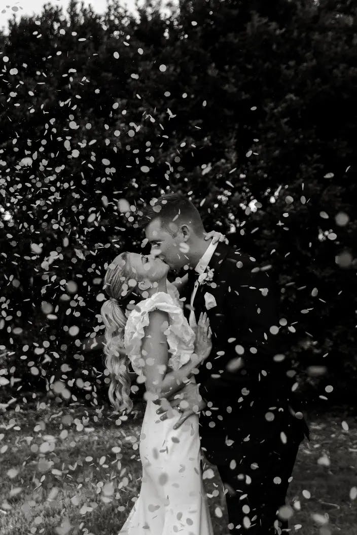 A bride and groom embrace and kiss outdoors amidst a shower of flower petals. The bride wears a white dress, and the groom is in a dark suit. They are surrounded by nature, with trees in the background. The image is in black and white.