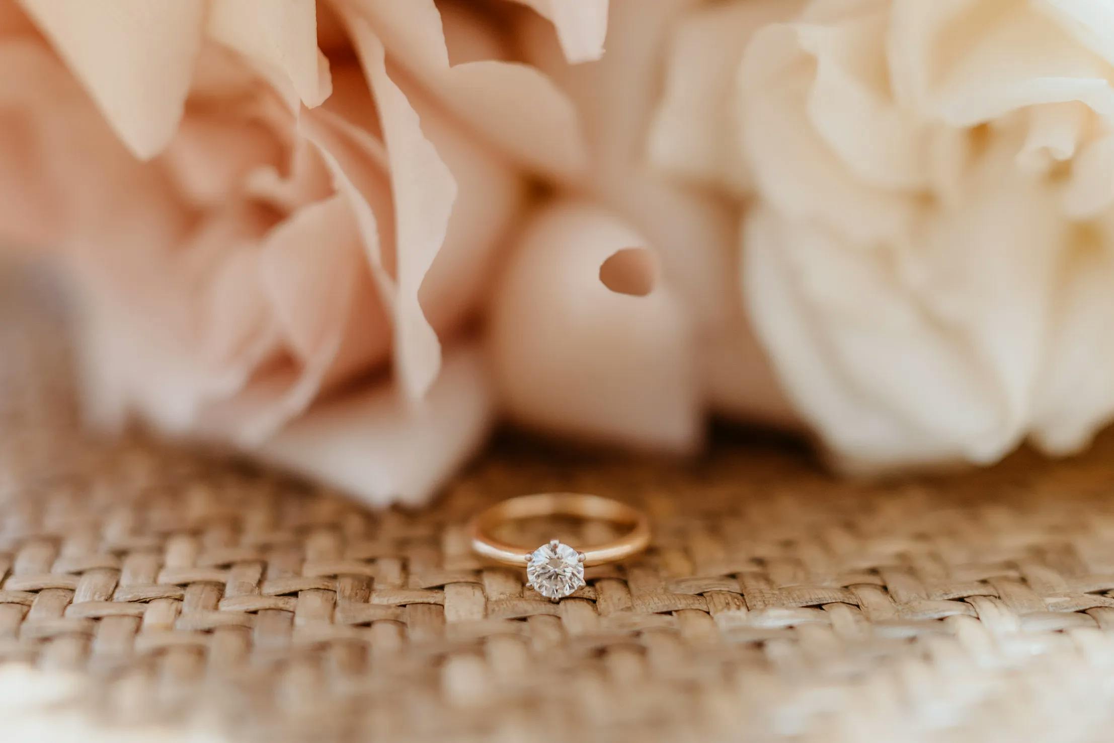 A gold engagement ring featuring a round diamond sits on a woven surface. Behind the ring, delicate blush and cream-colored flowers are slightly blurred, creating a soft, romantic background.