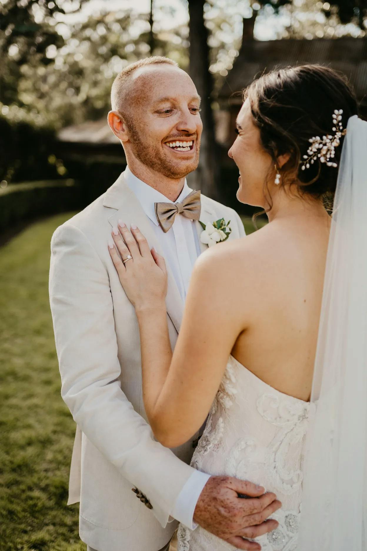 A bride and groom share a joyful moment outdoors on their wedding day. The groom, in a light-colored suit and bow tie, smiles at the bride, who is wearing a strapless gown and veil with sparkling hair accessories. They are holding each other lovingly, surrounded by greenery.
