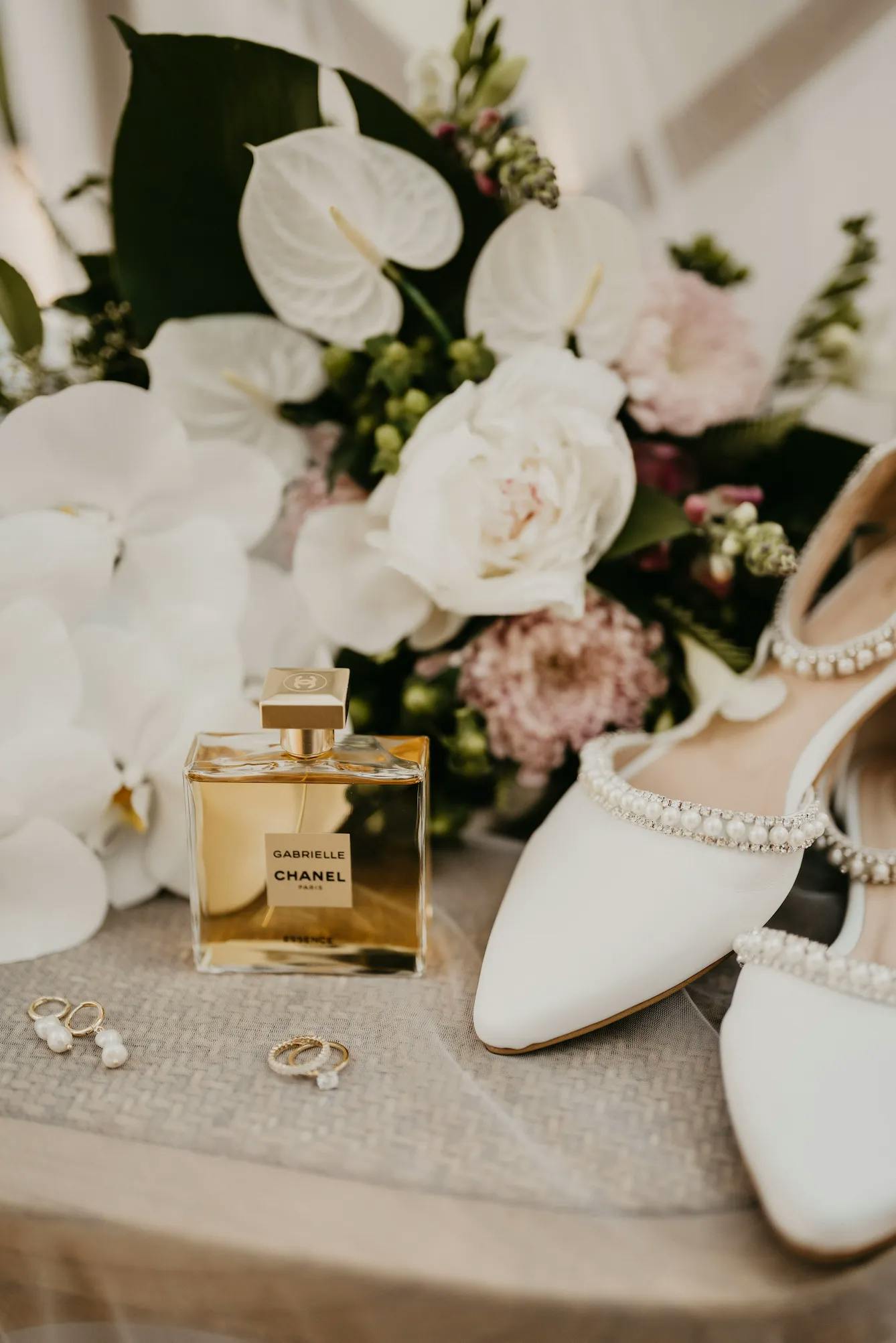 A photography setup features a Chanel perfume bottle, white bridal shoes adorned with pearls, a pair of pearl earrings, and a bouquet with a mix of white and pink flowers, all beautifully arranged on a textured surface.