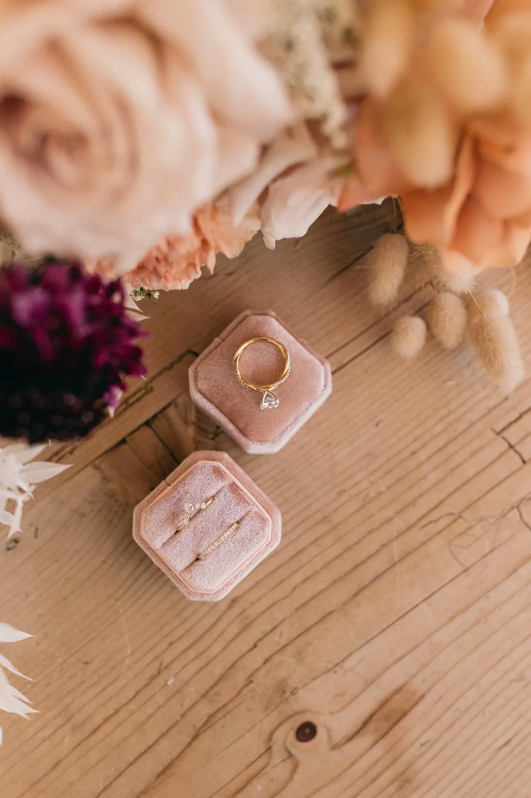 A pink velvet ring box with an open lid displaying a gold ring with a small diamond rests on a wooden table surrounded by various flowers in soft hues. A second, closed, matching pink velvet ring box is set beside it.