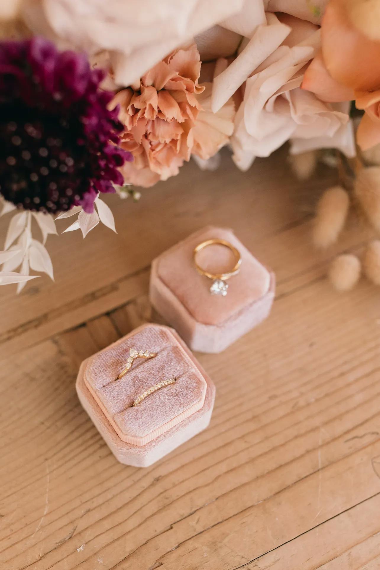 A wooden table holds two light pink velvet ring boxes. One box displays a gold ring with a single diamond, while the other shows two delicate gold rings. Above the boxes, a bouquet of flowers in shades of pink, peach, and dark purple is partially visible.