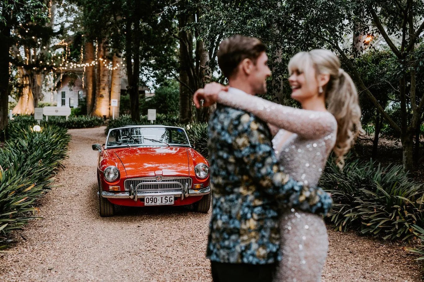 A couple in formal attire is happily embracing with a classic red car in the background. The car is parked on a gravel path lined with greenery, and fairy lights are strung between trees, creating a romantic atmosphere. The image is slightly blurred, focusing on the couple.