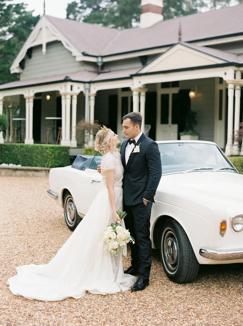 A bride and groom, dressed in a white gown and black suit respectively, stand next to a vintage white convertible car. The bride holds a bouquet of white flowers. They are positioned in front of a large, elegant house with a porch and peaked roofs.