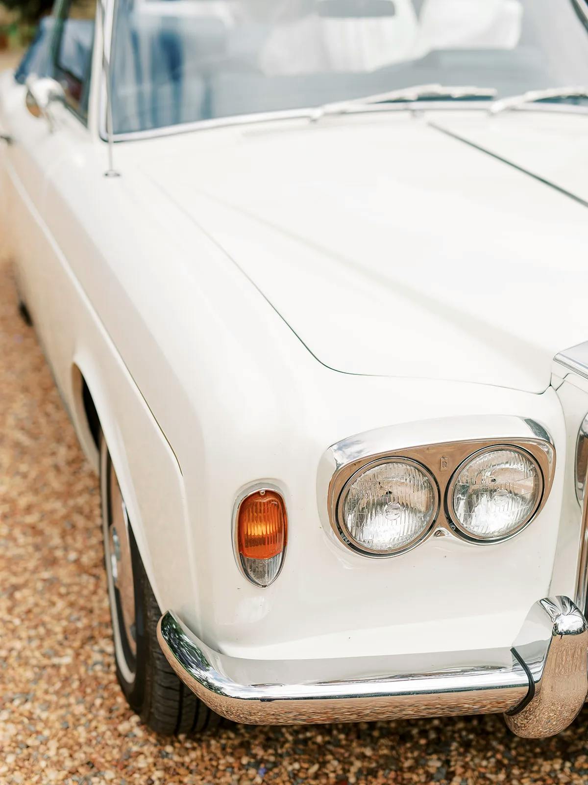 Close-up shot of the front left side of a vintage white car, showcasing the headlight, turn signal, and part of the chrome bumper. The car is parked on a gravel surface.