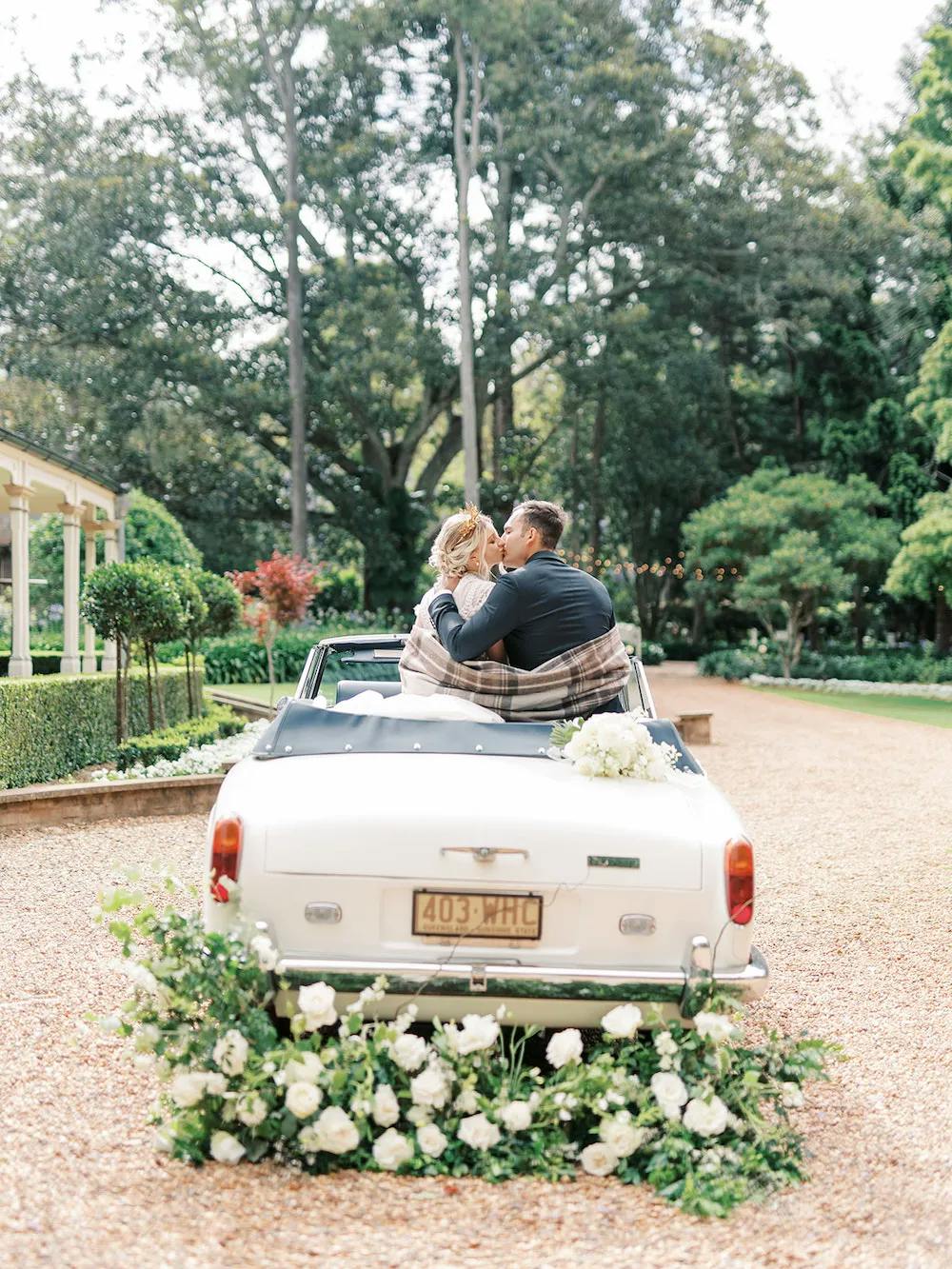 A couple sits on the back of a white convertible decorated with white flowers, sharing a romantic kiss. The car is parked on a gravel path in a lush garden, with tall trees and greenery in the background. The couple is wrapped in a blanket, adding to the intimate moment.