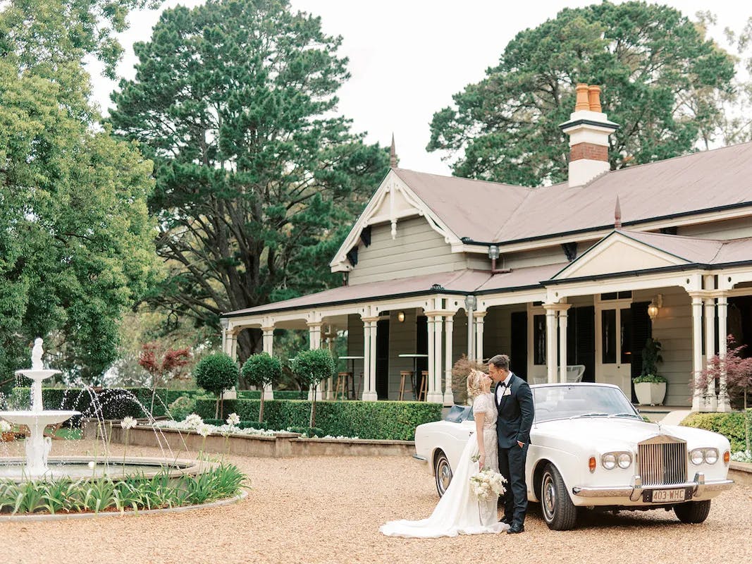 A bride and groom stand next to a classic white car in front of a large, elegant house surrounded by trees. The bride wears a white gown and the groom is in a dark suit. There is a white fountain to their left and the area is paved with gravel.