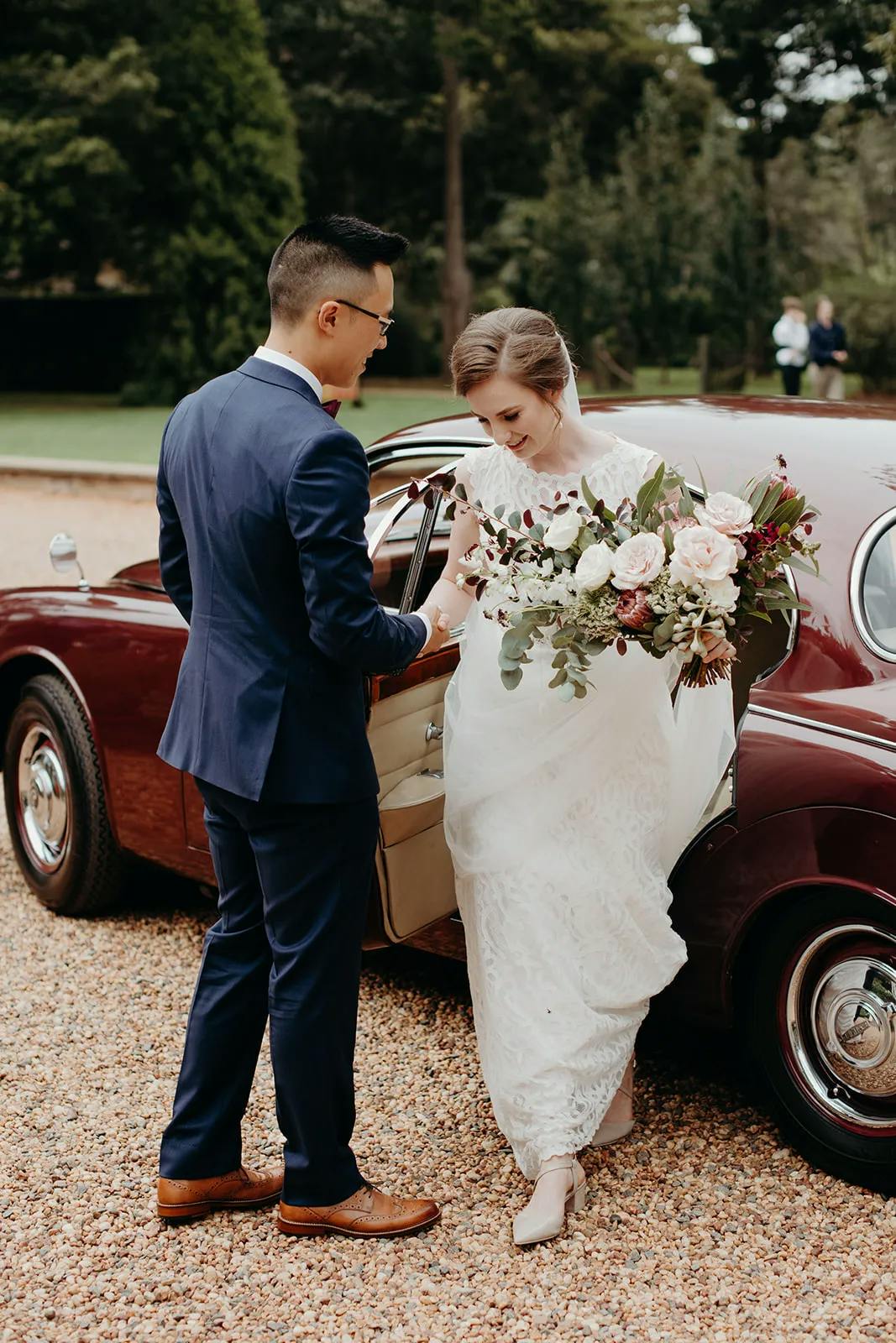 A bride in a white dress and groom in a blue suit stand next to a vintage maroon car. The groom holds the bride's bouquet of flowers as she steps out of the car. Trees and greenery are blurred in the background.