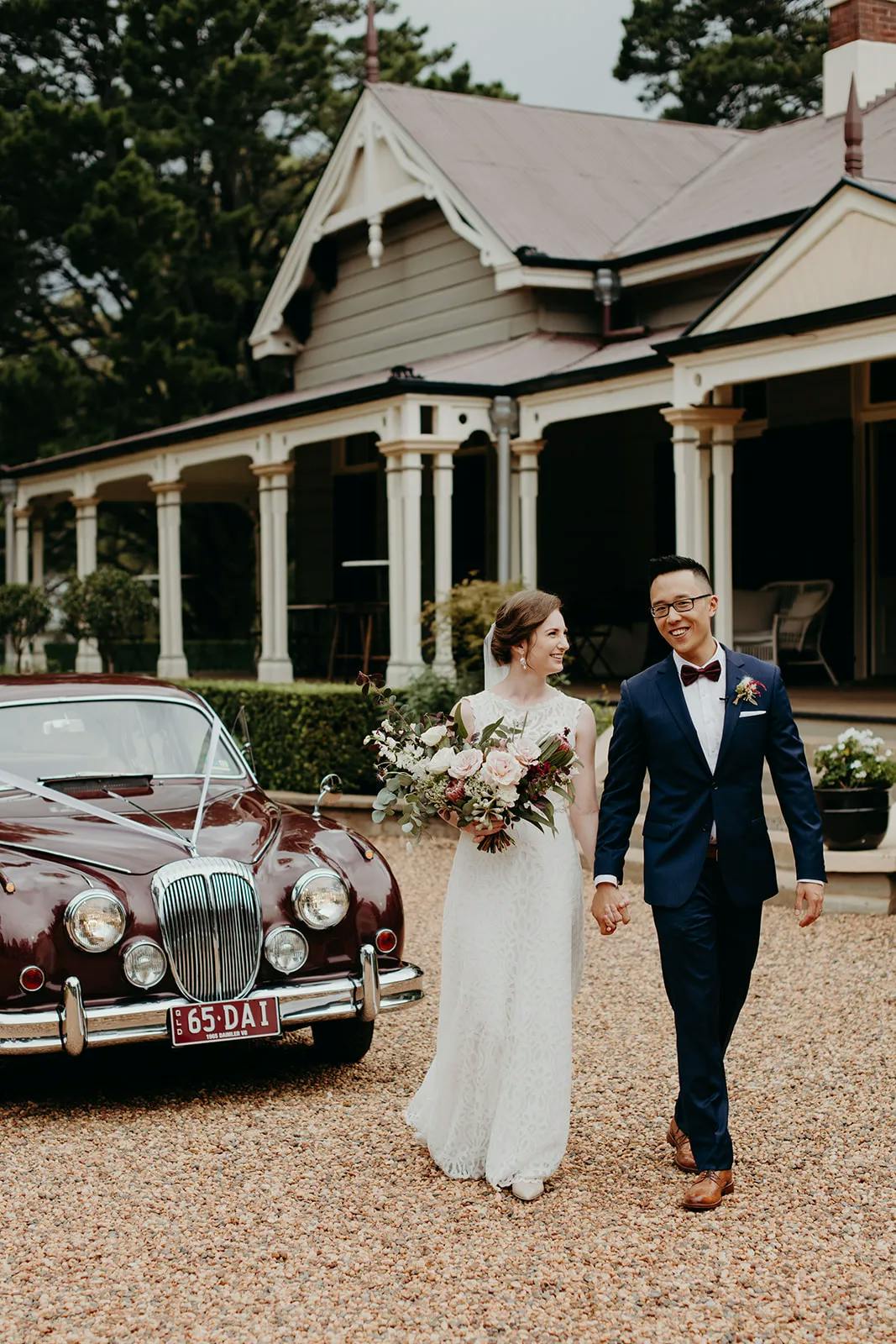A bride in a white dress holding a bouquet and a groom in a navy suit walk hand in hand in front of a vintage car and a large house with a covered porch and columns. They both appear happy, with the groom looking at the bride and smiling.