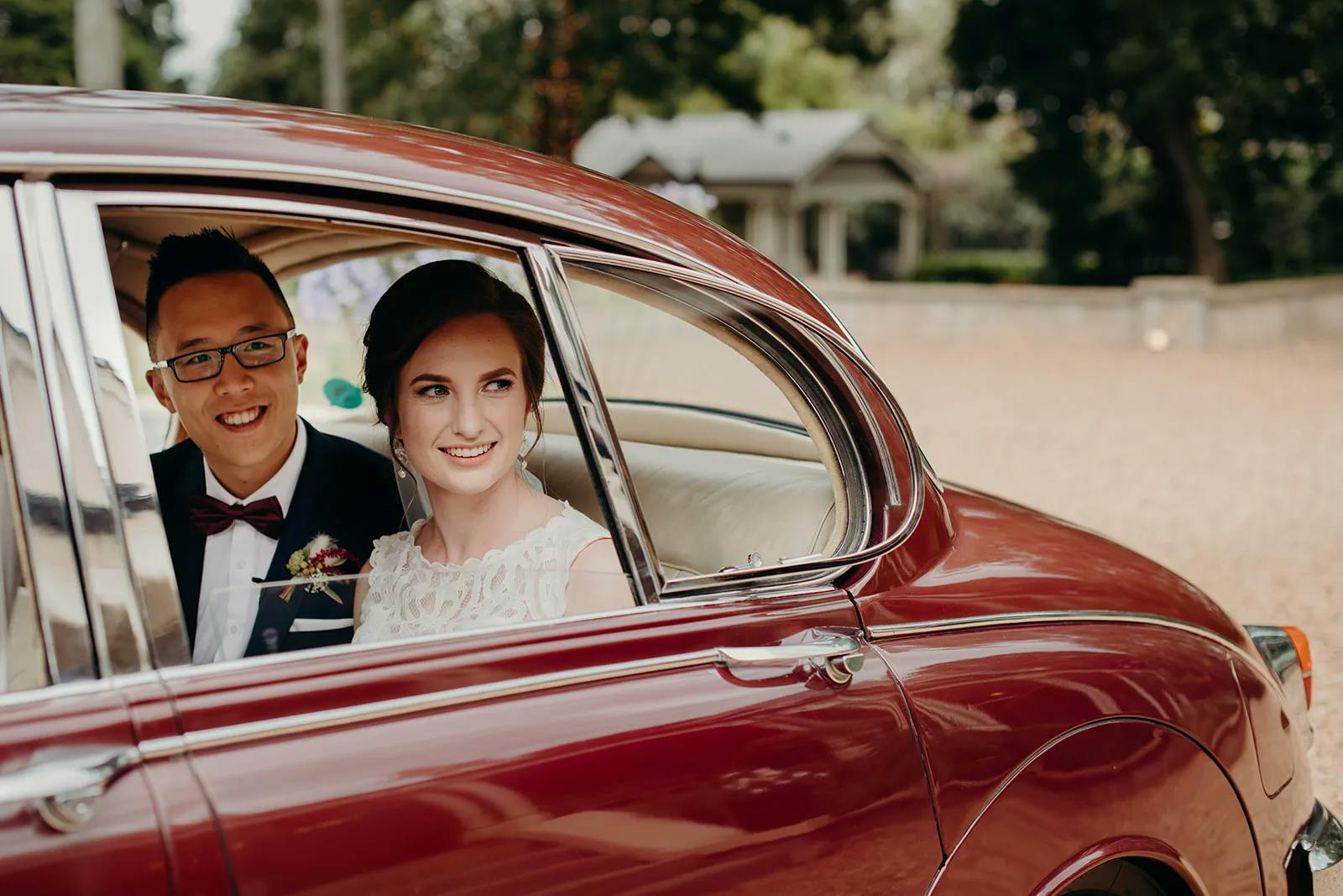 A smiling bride, wearing a white lace wedding dress, and a smiling groom, dressed in a tuxedo with a boutonniere, are sitting in the backseat of a vintage red car with the windows rolled down. They appear happy and excited, looking out the window. The background is an outdoor setting with trees and a building.