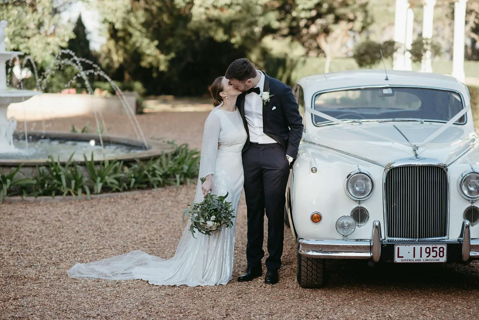 A bride in a white dress and a groom in a black suit share a kiss in front of a classic white car with a "L - 1958" license plate. They stand on a gravel path with a fountain and greenery in the background. The bride holds a bouquet of green foliage.