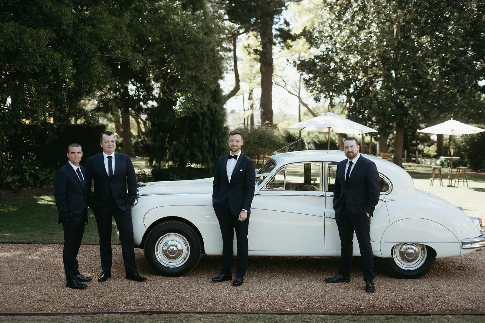 Four men dressed in formal suits stand in front of a vintage white car parked on a gravel path. Trees and a garden area with patio umbrellas are visible in the background. The men are posed casually, with two slightly leaning on the car.
