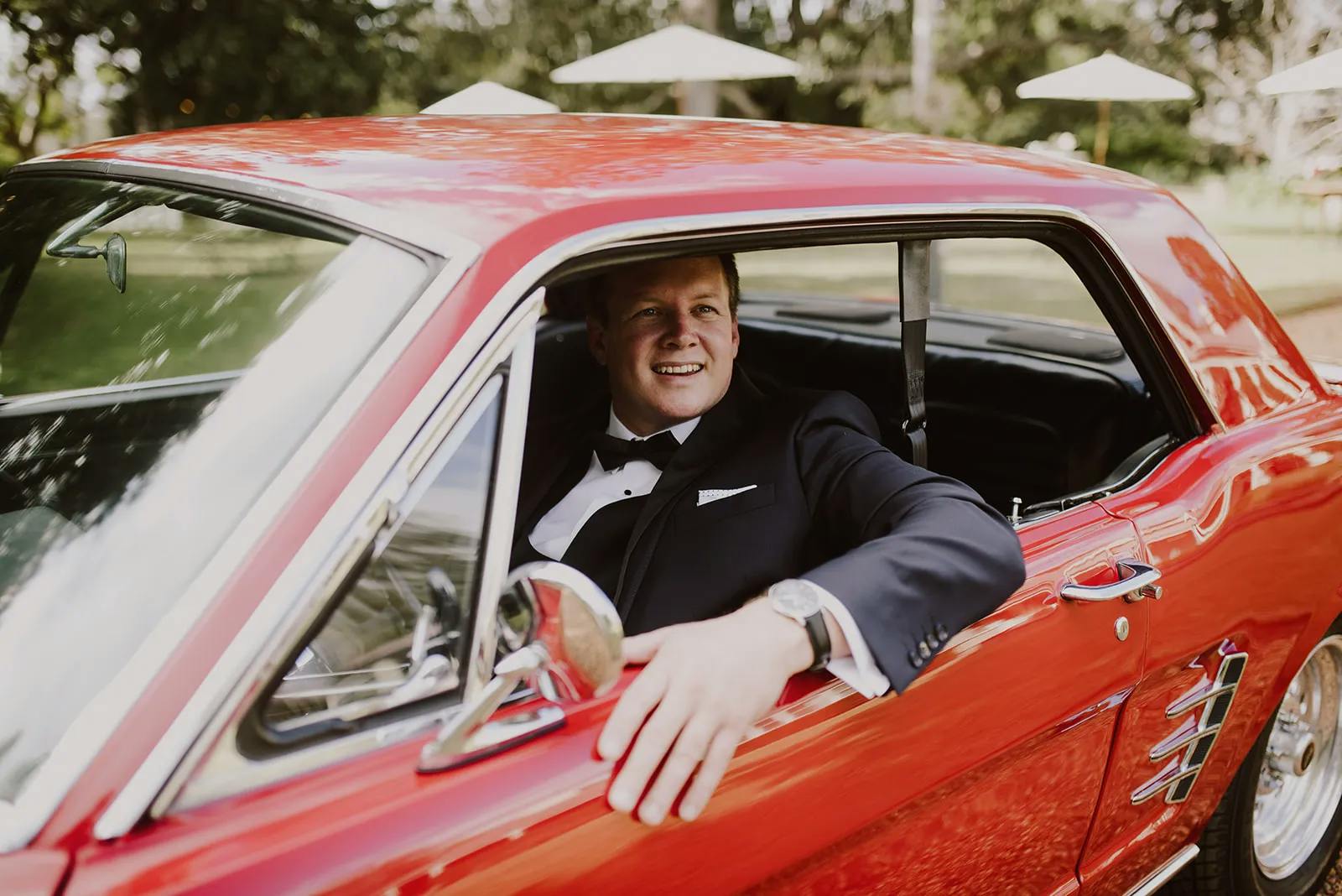 A man in a black tuxedo sits in the driver's seat of a classic red car, with the window rolled down and his arm resting on the ledge. He is smiling and looking out of the window. Sunlight filters through trees in the background.