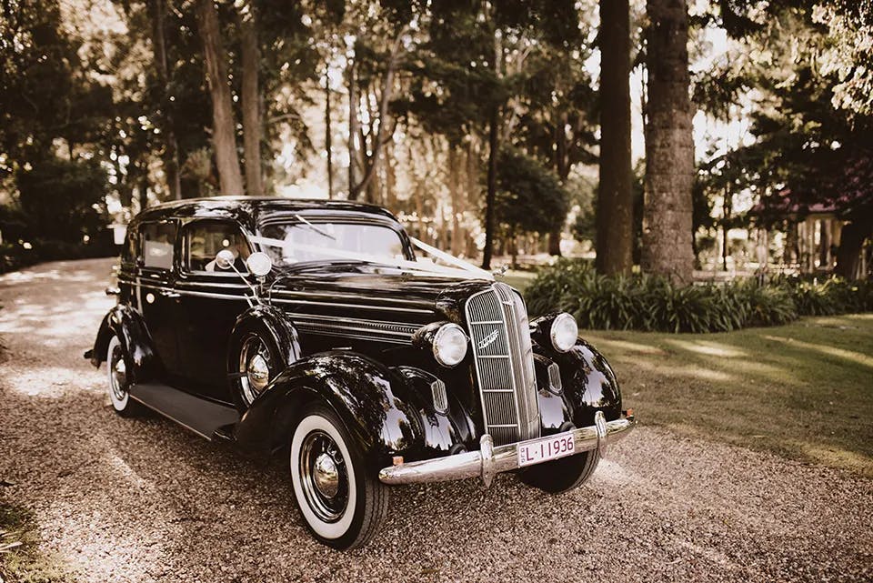 A vintage black car with white wall tires is parked on a gravel path in a wooded area. Tall trees and greenery surround the vehicle, and sunlight filters through the leaves, creating a nostalgic atmosphere. The car has a classic design with round headlights and a large grille.