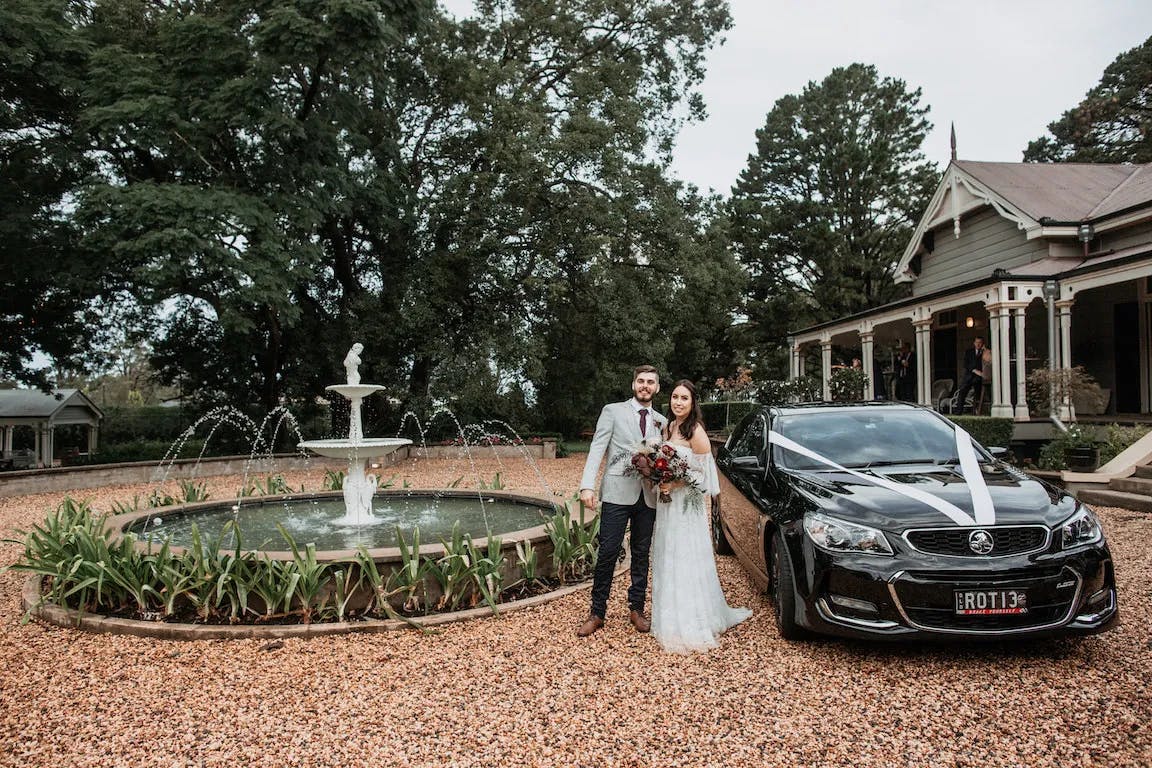 A newlywed couple stands beside a black, ribbon-adorned car on a gravel driveway. The groom wears a gray suit, and the bride wears a white dress and holds a bouquet. Behind them is a fountain and a large house with a porch, surrounded by greenery and trees.