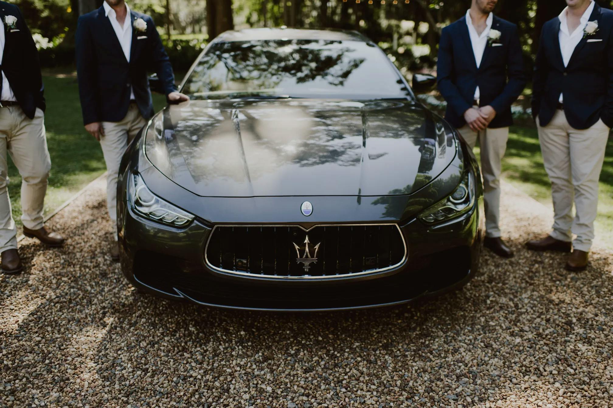 A sleek black Maserati sedan is parked on a gravel driveway, reflecting the surrounding trees and sunlight. Three men in dark blazers and light-colored pants stand on either side of the car, their faces out of frame.