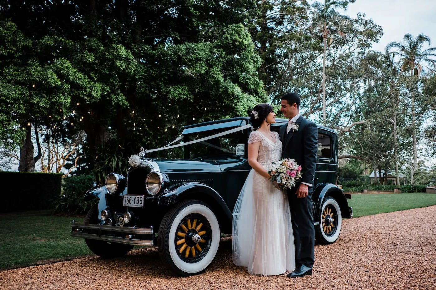A bride in a white dress and groom in a dark suit stand beside a vintage black car adorned with ribbons and flowers. They are outdoors in a lush, tree-filled setting with a gravel path. The bride holds a bouquet, and they look at each other lovingly.