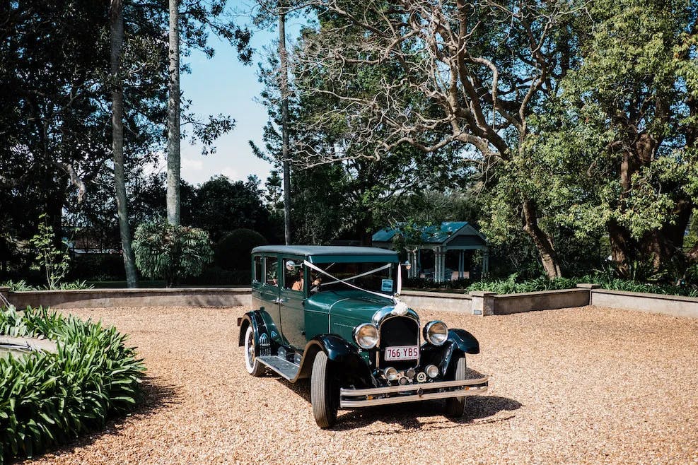 A vintage green car with white-walled tires is parked on a gravel driveway surrounded by lush greenery, trees, and a small structure in the background on a bright sunny day.