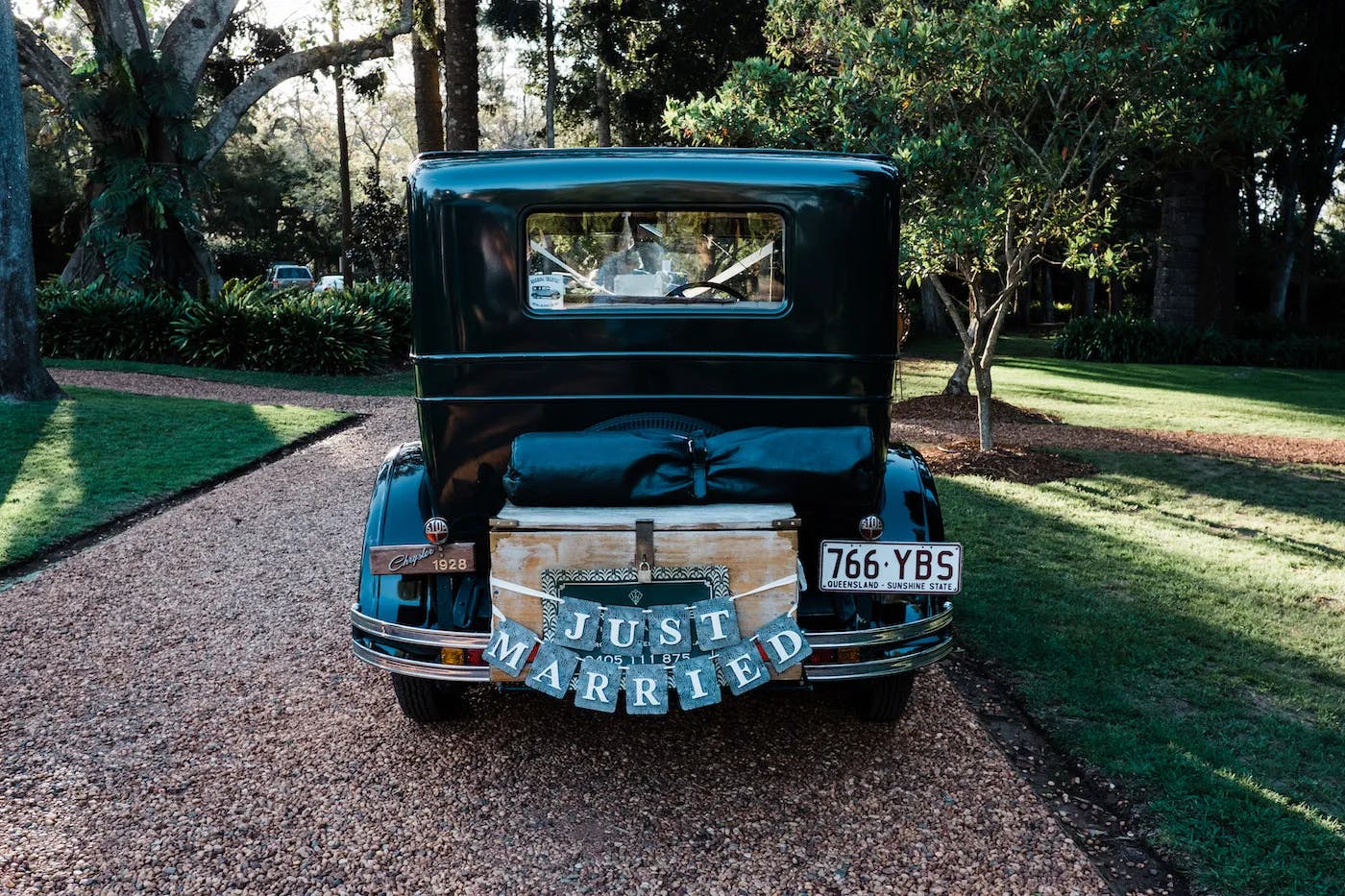 A vintage car with a "Just Married" sign on the back, adorned with small decorations, is parked on a gravel driveway. The surrounding area includes trees and neatly manicured grass, suggesting a serene, outdoor setting.