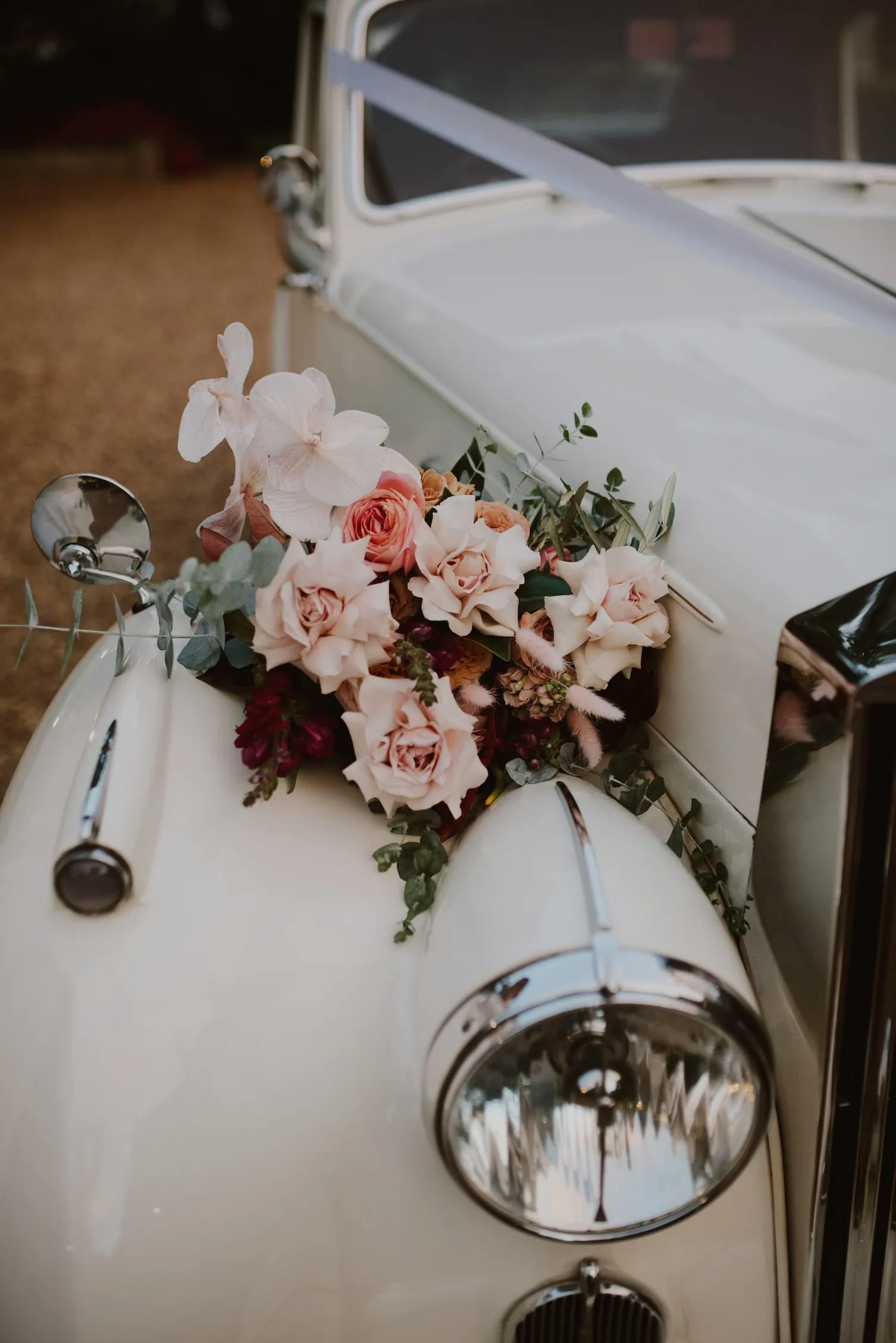 A vintage white car is adorned with a bouquet of pink and white roses along with greenery on the front hood, near the headlight. The car has a satin ribbon attached, suggesting it is decorated for a wedding or special event.