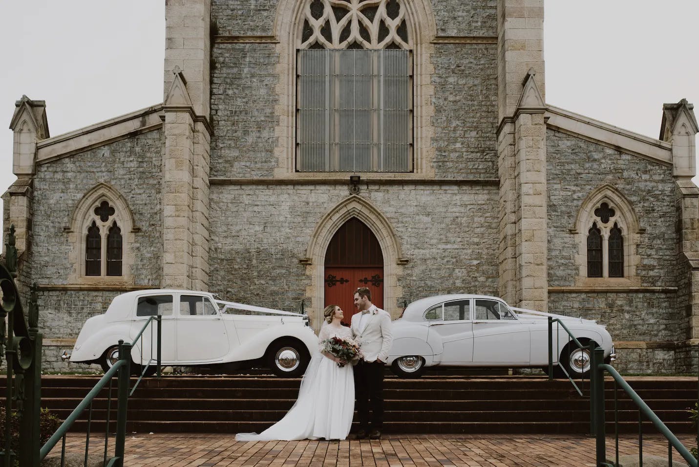 A bride and groom stand in front of an old stone church with large arched windows and a tall door. The bride wears a white gown and holds a bouquet, while the groom wears a light-colored suit. Two vintage white cars are parked on either side of the stairs.
