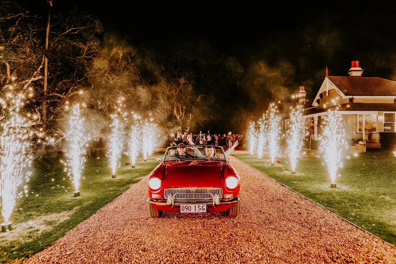 A red convertible car drives down a gravel path lined with sparklers on both sides, creating a festive atmosphere. The path leads to a house in the background, with people visible near the entrance. The scene is set at night, and the sparklers illuminate the surroundings.