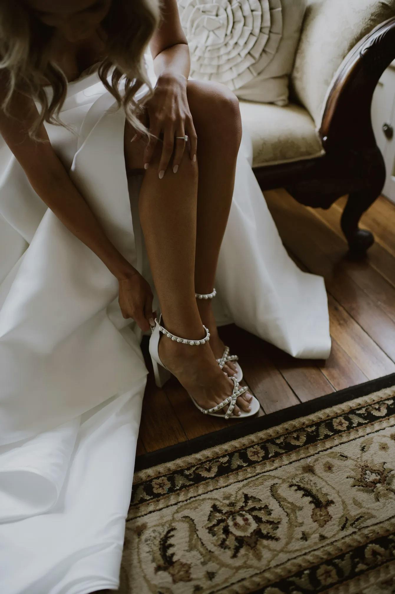 A woman in a white dress is sitting on an upholstered chair and adjusting the strap of one of her pearl-embellished high-heeled sandals. The scene includes a patterned rug and wooden flooring.