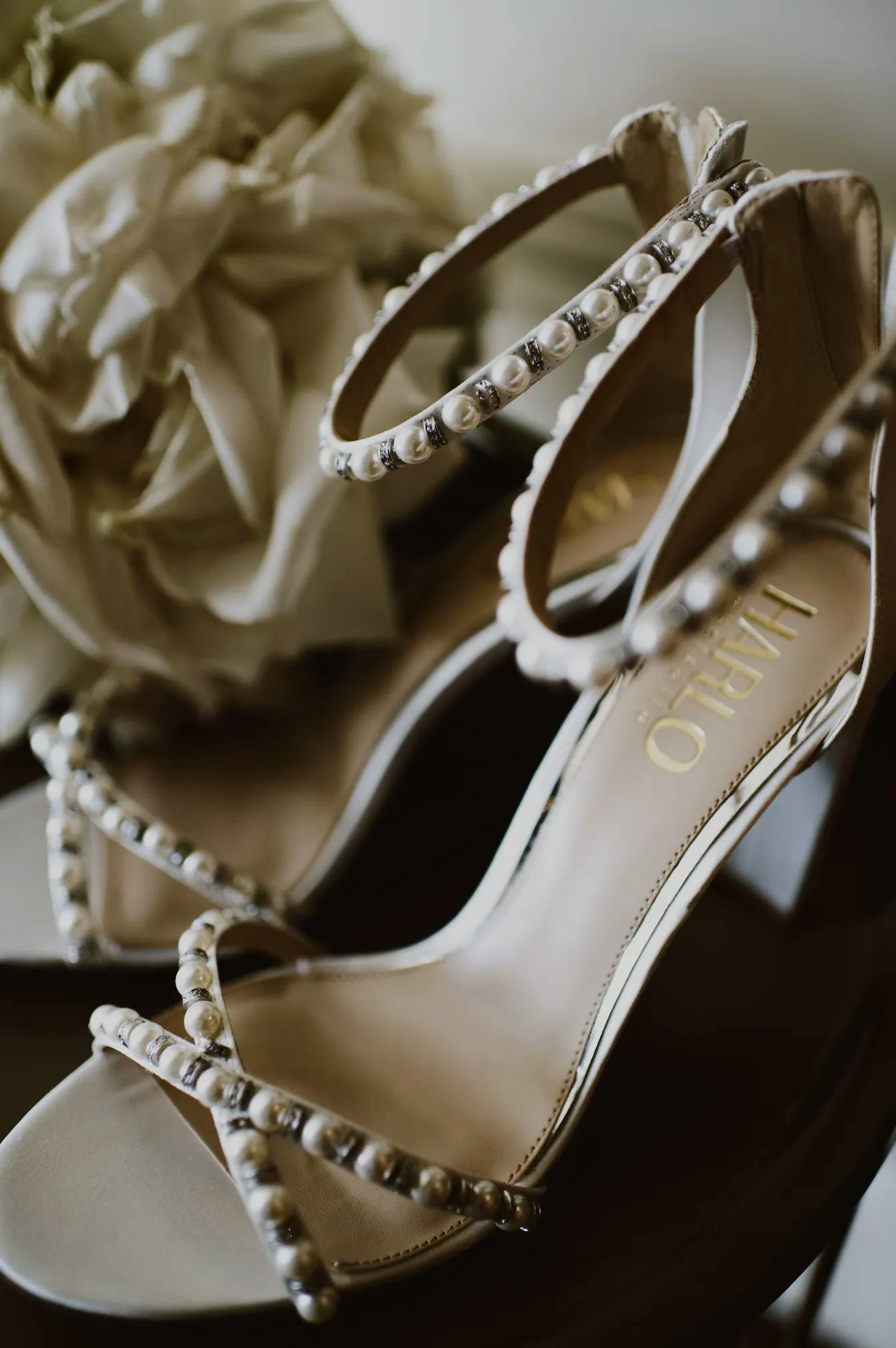 Close-up of a pair of elegant open-toe high-heeled shoes decorated with pearls, placed on a dark surface. A bouquet of white flowers is softly blurred in the background, providing a delicate and sophisticated ambiance.