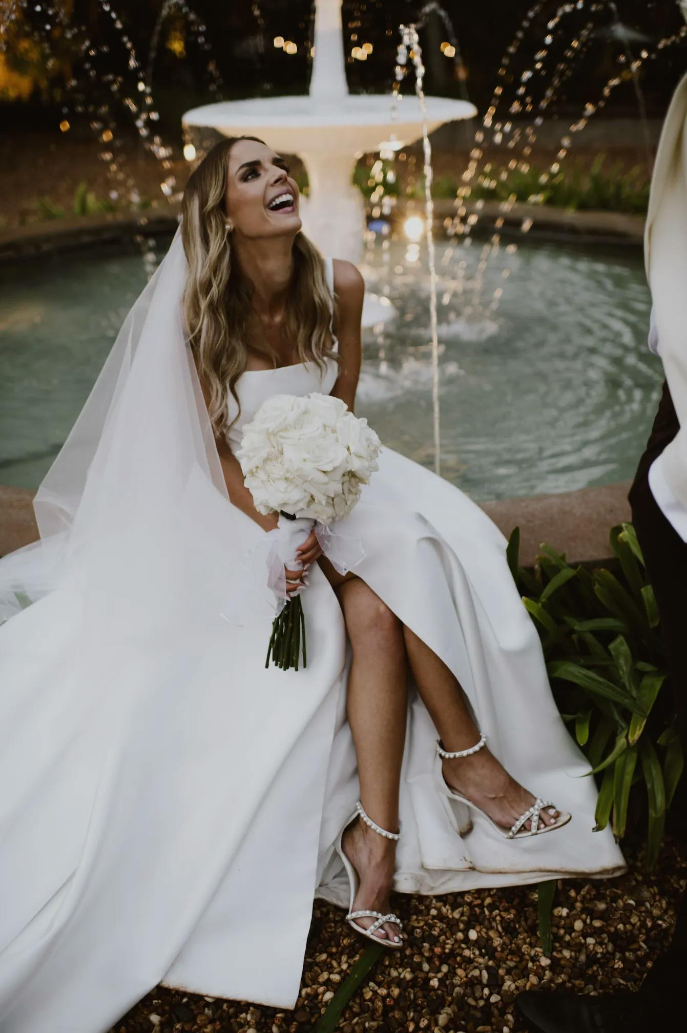 A bride in a white gown and veil sits by a fountain, holding a bouquet of white flowers. She smiles joyfully while wearing strappy heels decorated with pearls. The background features water spraying from the fountain and lush greenery.