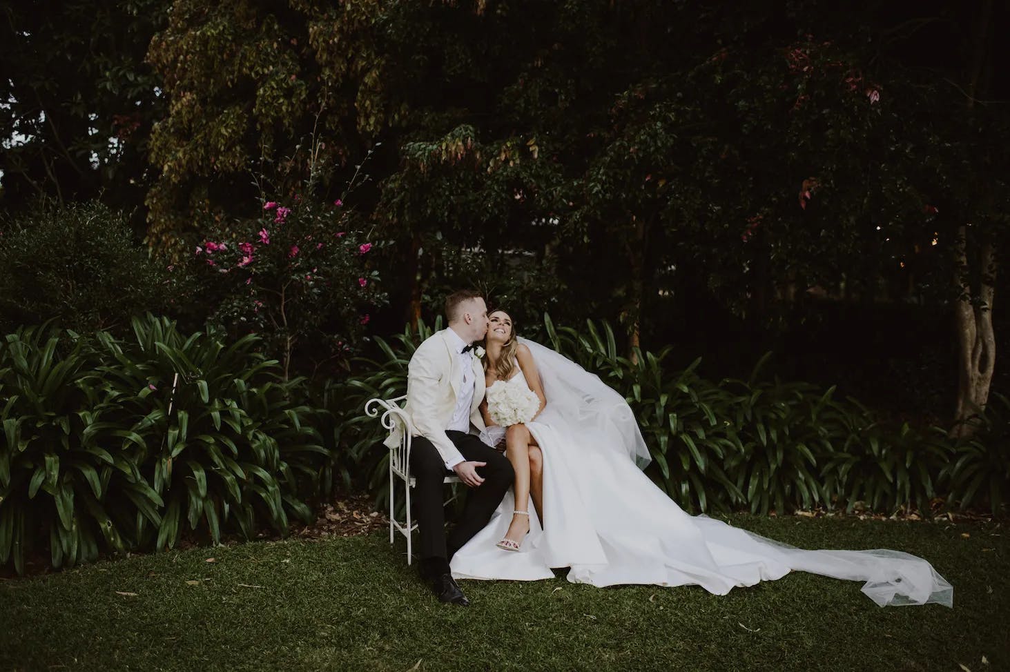 A bride and groom sit on a white bench in a lush, green garden. The groom, in a white jacket and black pants, kisses the bride, who is in a long white gown with a veil. They are surrounded by plants and flowers, enjoying a serene and intimate moment.